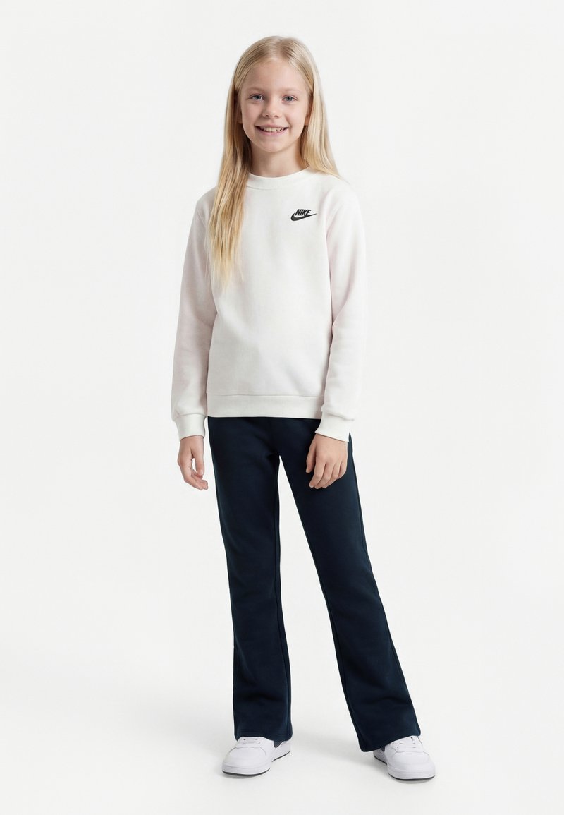 Ragazza giovane con lunghi capelli biondi, indossa una felpa bianca Nike, pantaloni neri e sneakers bianche, in piedi e sorridente.