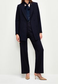 Blazer doppiopetto blu navy su una camicetta abbinata, abbinato a pantaloni su misura e scarpe con tacco nude, con una cravatta a fiocco di raso.