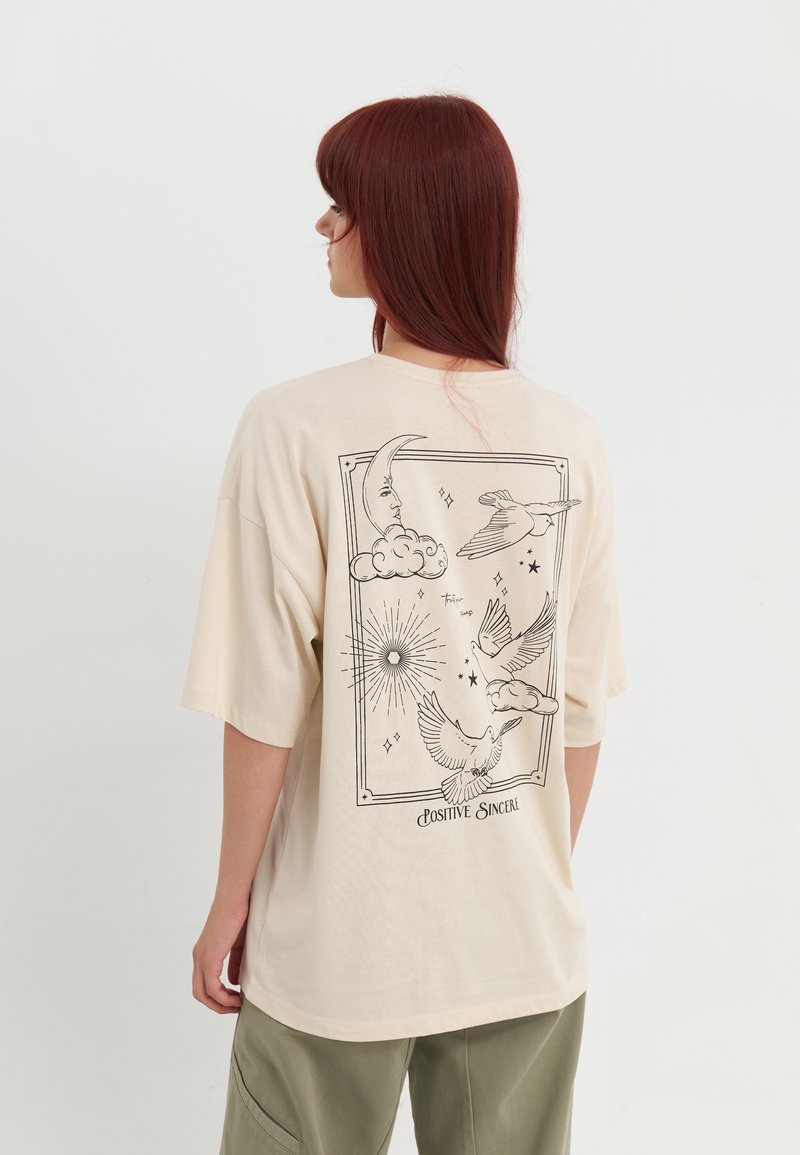 T-shirt oversize beige à manches courtes ; affiche des illustrations en ligne noire d'éléments célestes et d'oiseaux au dos ; matière douce en coton.