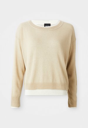 Suéter beige de manga larga y cuello redondo, con puños y dobladillo acanalados, superpuesto a una prenda blanca, con una etiqueta negra en el cuello.