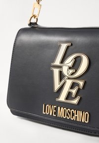 Bolsa de mão de couro preto com ferragens douradas. Apresenta um logo em destaque com "LOVE" em tamanhos de fonte variados e "LOVE MOSCHINO" estampado na parte inferior.