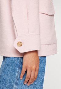 Main dans la poche d’un jean en denim bleu portant une manche de manteau rose clair avec un bouton marron au poignet.