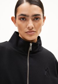 Schwarze Zip-Jacke aus weichem Stoff, mit einem hohen Kragen und gesticktem Logodetail auf der Brust. Silberner Reißverschluss-Akzent.