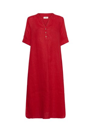 Žena s dlhými vlnitými vlasmi nosí červené midi šaty na gombíky a hnedé sandále na platforme, stojaca so zloženými rukami na bielom pozadí.