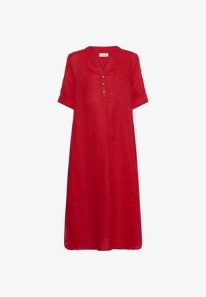 Žena s dlhými vlnitými vlasmi nosí červené midi šaty na gombíky a hnedé sandále na platforme, stojaca so zloženými rukami na bielom pozadí.