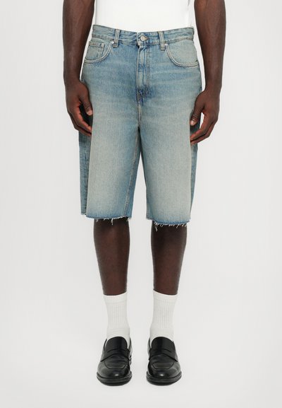 Un short en denim bleu clair à coupe droite, avec un ourlet brut, des poches à l'avant et une légère décoloration. Associé à des chaussettes blanches et des mocassins noirs.