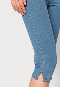 Světle modré džínové capri kalhoty s zužujícím se střihem, s neupraveným lemem se štěrbinou, viditelným šitím a hladkou texturou.