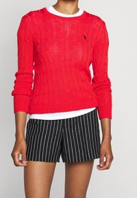 Röd kabelstickad tröja med långa ärmar, rund halsringning och liten logotypdetalj; bärs över en vit topp och matchas med svart randiga shorts.