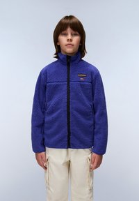 Giacca in pile viola con colletto alto, chiusura con zip e superficie strutturata; presenta un piccolo logo sul petto. Indossata con pantaloni cargo beige.
