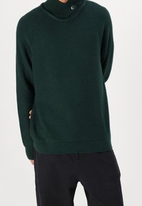 Dunkelgrüner Pullover mit hohem Kragen und Knopfdetails. Hergestellt aus einem weichen, strukturierten Stoff, mit gerippten Bündchen und Saum.