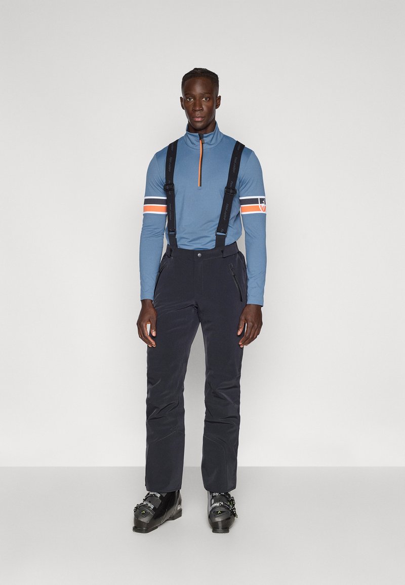 Blaues Langarm-Zip-Up-Shirt mit orangefarbenen Akzenten, kombiniert mit schwarzen Skihosen mit verstellbaren Trägern und Reißverschlüssen. Schwarze Skischuhe sind ebenfalls enthalten.