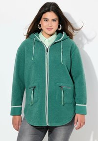 Veste en polaire verte avec capuche, fermeture éclair frontale, deux poches zippées et bord contrastant blanc sur les poignets et la fermeture éclair. Texture douce.