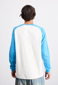 Chemise à manches longues avec un corps blanc et des manches raglan bleu ciel. Texture lisse avec des poignets côtelés et un col rond.