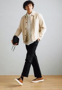 Chaqueta ligera beige sobre una camiseta blanca, pantalones negros a medida y zapatillas marrones. Sosteniendo un pequeño bolso negro, con un fondo sencillo.