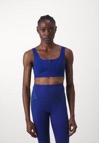 Sinine spordinärk laiemate rihmadega, ees lukuga ja perforatsioonidega detailideks. Ühendatud samas värvitoonis kõrge vöökohtaga retuusidega. Sile tekstuur.