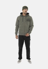 Sudadera verde con cremallera y bolsillo delantero, combinada con pantalones negros y zapatillas de cuadros. Los accesorios incluyen un gorro verde a juego.