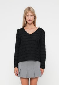 Zwarte langslavige top met een gestructureerd, golvend patroon en een V-hals, gecombineerd met een grijze geplooide rok, die een modern, casual ontwerp laat zien.