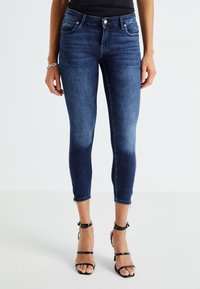 Jeans de mezclilla azul oscuro ajustados con una cintura de talle medio, longitud cropped por encima del tobillo y detalles con cremallera en el dobladillo. Textura suave con un ligero desvanecimiento.