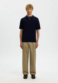 Jeune homme aux cheveux blonds courts portant une chemise bleu marine à col zippé à manches courtes, un pantalon beige à jambes larges et des chaussures marron foncé, debout.