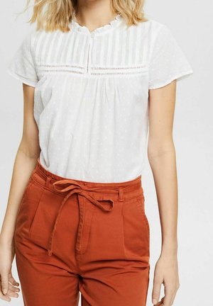 Kvinde iført hvid kortærmet bluse med blonde detaljer og højtaljede bukser i rustfarve, bundet med et stofbælte.
