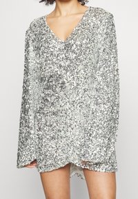 Stříbrný top se štrasovými flitry, s výstřihem do V, dlouhými rukávy a volným střihem. Textura je lesklá, s nepravidelnými stříbrnými a iridescentními vzory.