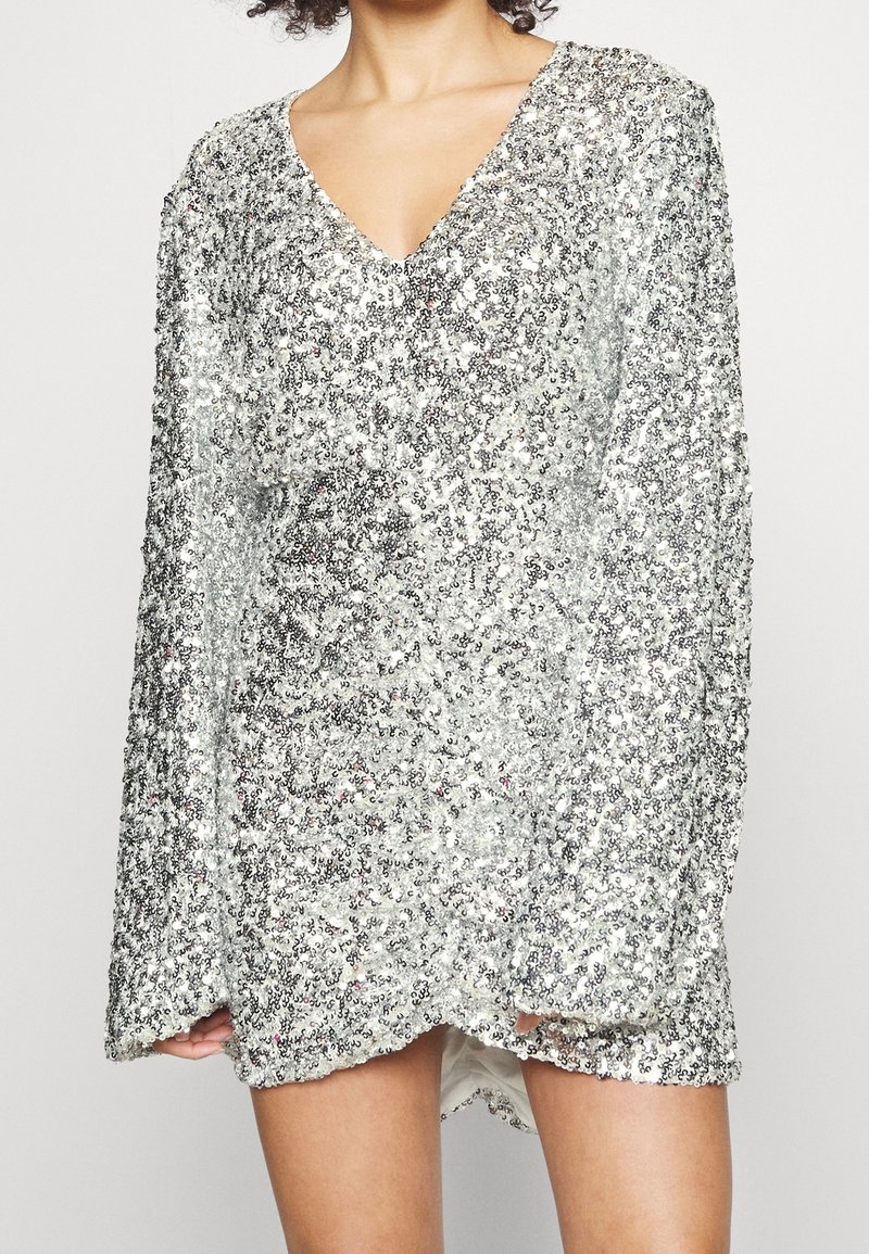 Stříbrný top se štrasovými flitry, s výstřihem do V, dlouhými rukávy a volným střihem. Textura je lesklá, s nepravidelnými stříbrnými a iridescentními vzory.