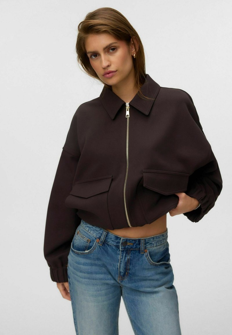 Jeune femme portant une veste courte marron foncé avec de grandes poches et un jean bleu, posant avec une main dans la poche sur un fond uni.