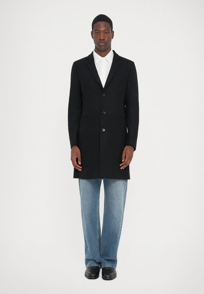Schwarzer Wollmantel mit tailliertem Design, einreihig, fünf Knöpfe, zwei seitliche Taschen, getragen über einem weißen Hemd und blauen Jeans.