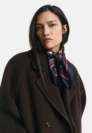 Femme jeune avec de longs cheveux noirs portant un manteau marron foncé et une écharpe florale bleu marine nouée autour du cou sur un fond blanc.