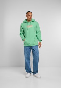 Miętowa bluza z kapturem z mieszanki bawełny z przednią kieszenią kangurką i grafiką na piersi, w zestawieniu z jasnoniebieskimi dżinsami oraz białymi trampkami.