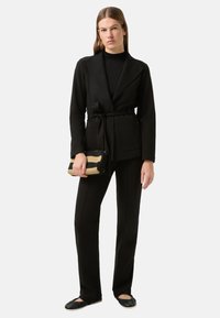 Schwarzer, maßgeschneiderter Blazer mit Gürtel, kombiniert mit schwarzen, eng anliegenden Hosen und schwarzen Ballerinas. Hält eine zweifarbige Handtasche mit gestreiftem Muster.