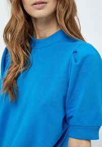 En ljusblå sweatshirt med rund halsringning, korta puffärmar och ribbade muddkanter. Mjuk tygtextur med en slät yta.