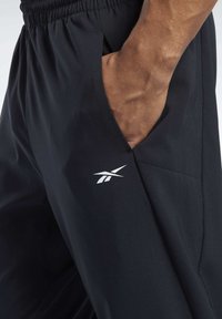 Černé sportovní šortky s elastickým pasem, s boční kapsou a bílým logem Reebok poblíž pasu. Hladká, lehká tkanina.