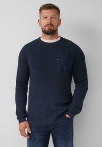 Dunkelblauer gestrickter Pullover mit gerippter Textur, Raglanärmeln und einer Brusttasche mit Knopfdetail. Getragen über blauen Jeans.