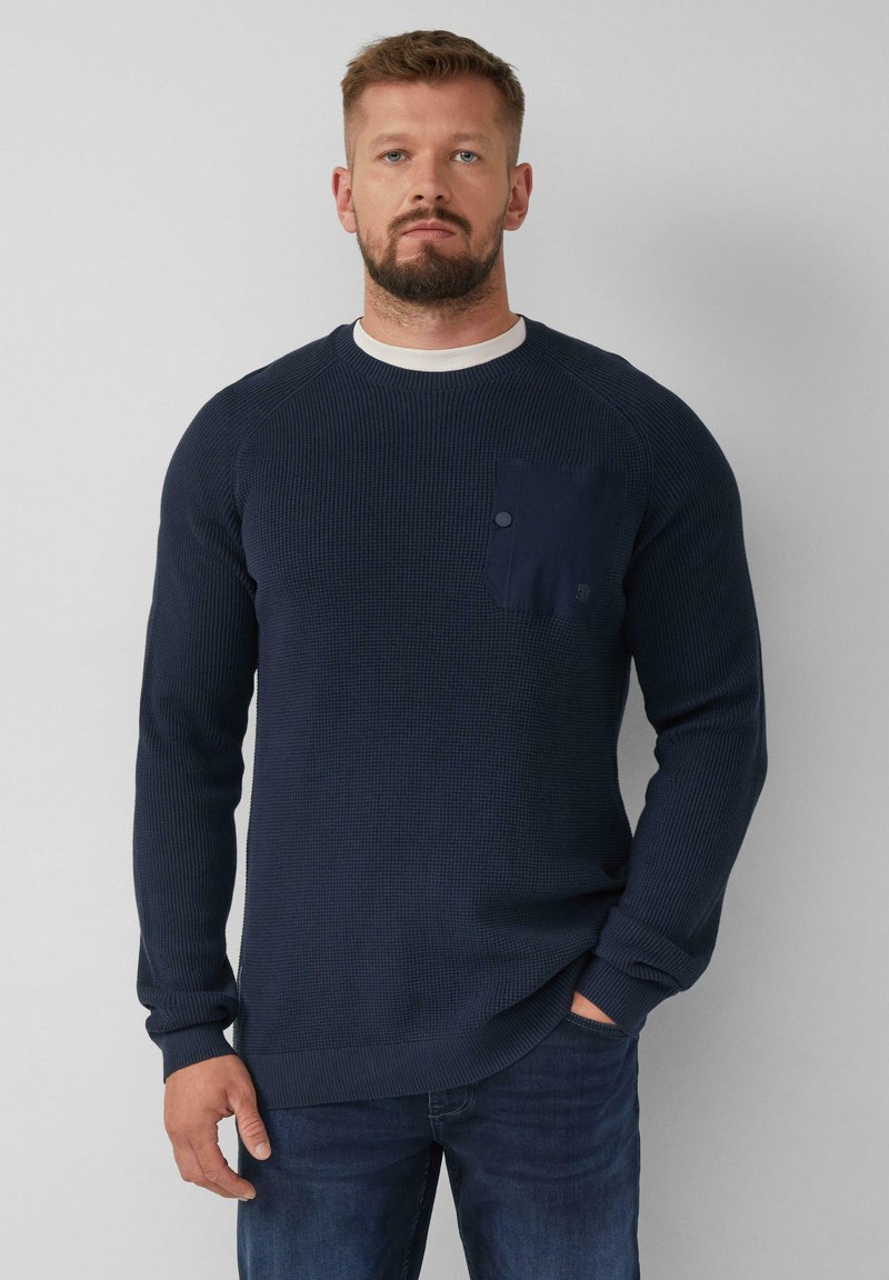 Dunkelblauer gestrickter Pullover mit gerippter Textur, Raglanärmeln und einer Brusttasche mit Knopfdetail. Getragen über blauen Jeans.