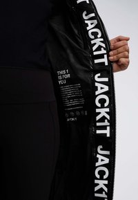 Una mano tiene aperto un giubbotto nero con la scritta bianca "JACKIT" lungo la zip e un testo incentrato sulla sostenibilità all'interno della foderatura del giubbotto.