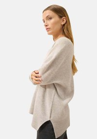 Lichtgrijze gebreide trui met een losse pasvorm, verlaagde schouders, zijsplitten en ribbeltextuur. Gecombineerd met donkere jeans op een witte achtergrond.