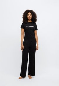 Tricou negru cu logo-ul "KARL LAGERFELD" și pantaloni negri cu picioare largi, ambele realizate dintr-un material neted, purtate de un model desculț.