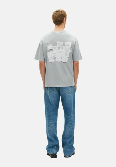 Camiseta de algodón gris de manga corta, con notas escritas a mano en papel pegadas en la parte trasera. Lleva puesta un pantalón de mezclilla azul claro.