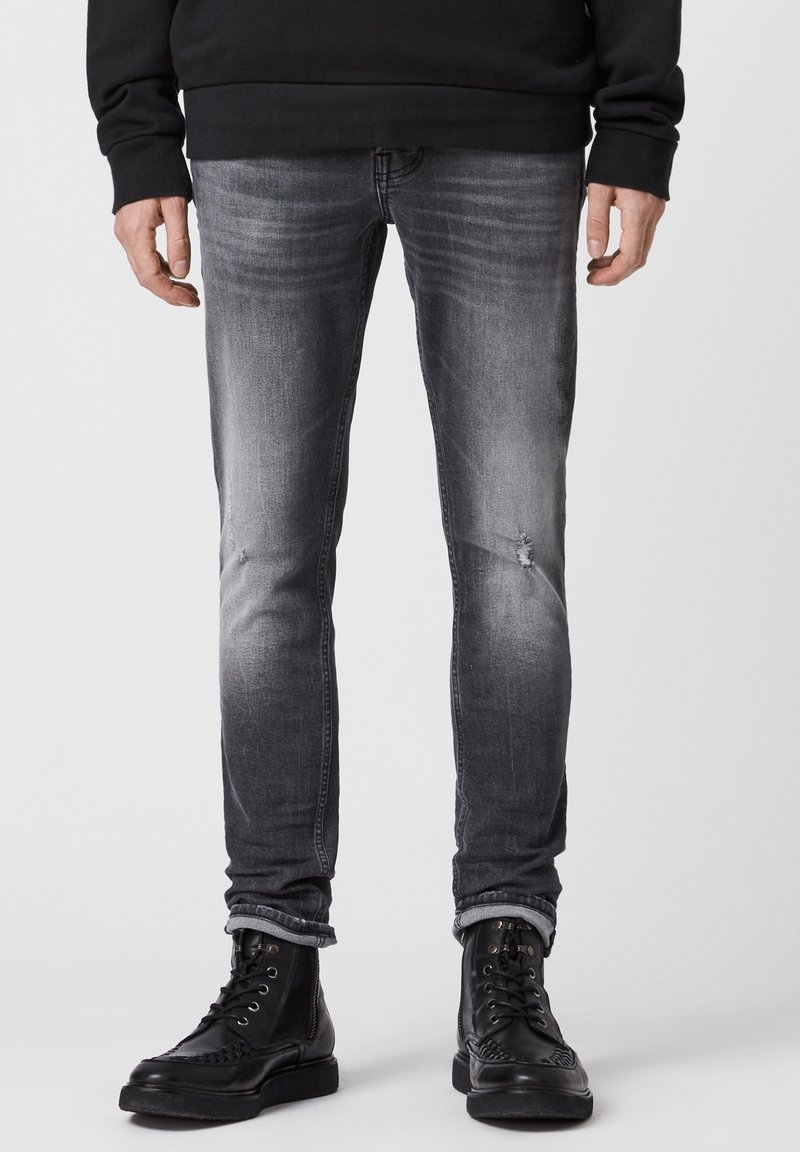 Jean en denim gris coupe slim avec une finition délavée, présentant une légère usure aux genoux, associé à des bottines noires à lacets.