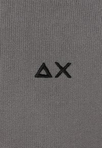 Tessuto lavorato a maglia grigio con un triangolo ricamato nero e simbolo 'X', caratterizzato da una fine costina su tutta la superficie.