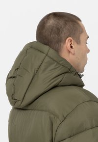 Alyvuogių žalias pūkinis švarkas su gobtuvu, turintis quilted tekstūrą ir aukštą apykaklę. Medžiaga atrodo lengva ir atspari drėgmei.