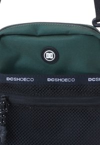 Zelená látková taška s čiernou sieťovanou dolnou časťou, ktorá má okrúhly logo a zipsové uzatvorenie. Popruh je nastaviteľný a má kovové doplnky.