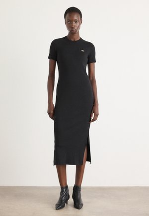 Robe en tricot noire à manches courtes, slim fit, décolleté rond, fente sur le côté et petit logo sur la poitrine. Associée à des bottines noires.