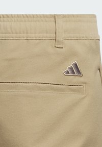 Pantalones cortos caquí con un bolsillo trasero horizontal, banda elástica en la cintura y un logo de Adidas bordado en dorado en el lateral.