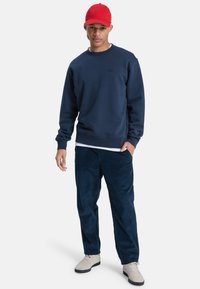 Marineblauwe sweatshirt met ronde hals, zachte textuur en klein logo; gecombineerd met marineblauwe corduroypantalon en lichtgrijze schoenen. Rode pet op het hoofd.