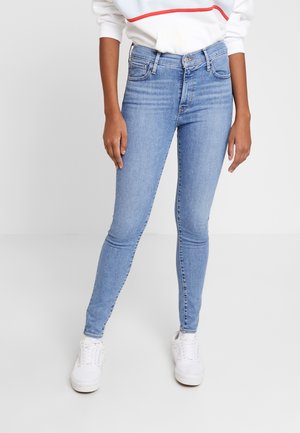 Personne portant un jean skinny bleu clair, des baskets blanches et un sweat-shirt blanc avec une bande rouge, debout devant un fond blanc.