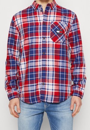 Mężczyzna noszący czerwoną, niebieską i białą koszulę w kratę na guziki z kieszenią na piersi z napisem Tommy Jeans, w połączeniu z jasnoniebieskimi dżinsami.