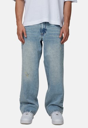 Mężczyzna noszący jasnoniebieskie jeansy o prostych nogawkach z delikatnym przetarciem, białe trampki oraz białą krótką koszulę z krótkim rękawem.