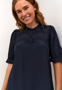 Temno modra bluzica s kratkimi naborki, z čipkastim panelom čez prsi, gumbnim zapiranjem spredaj in gladko teksturo.